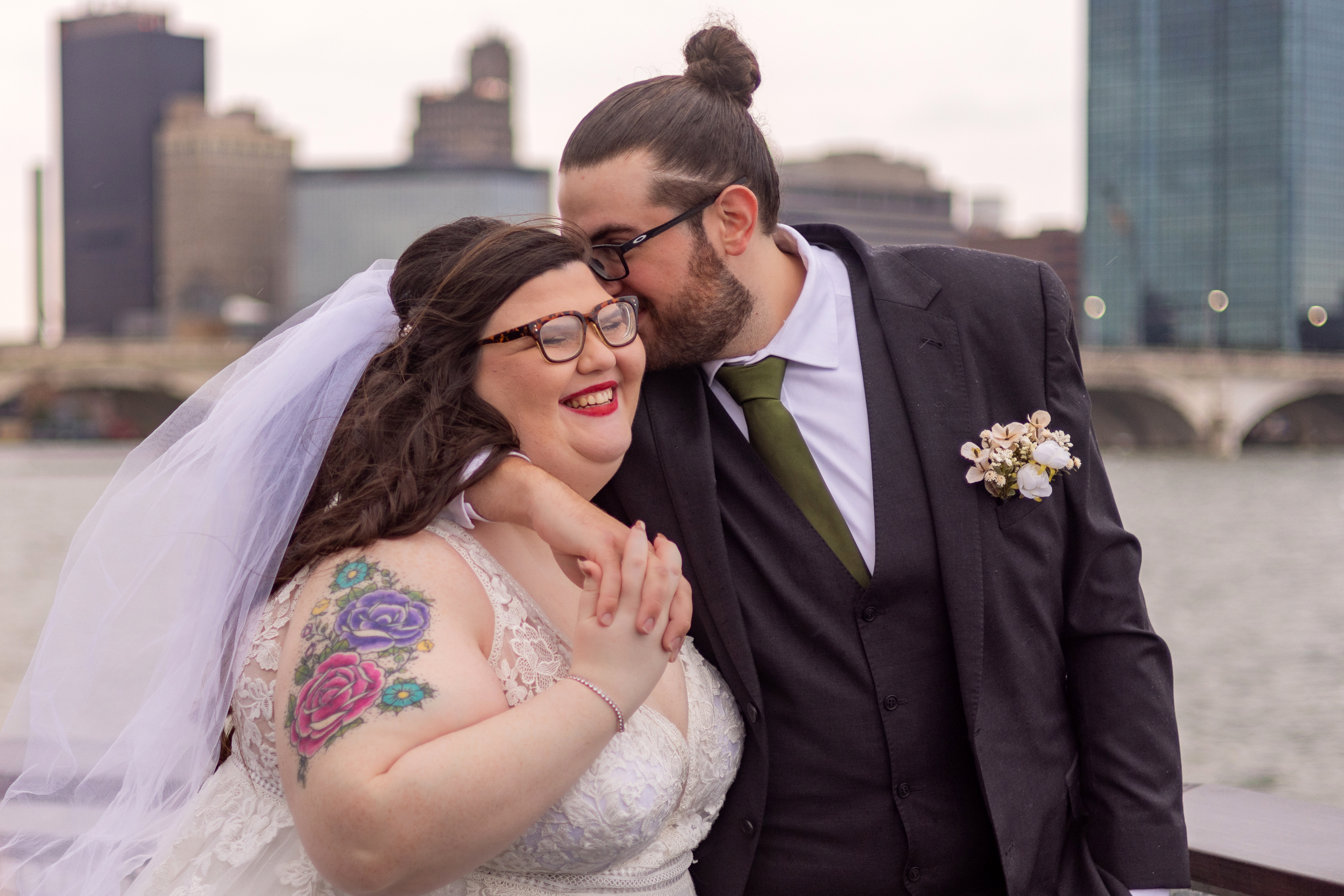 groom kissing bride on cheek with city skyline in background