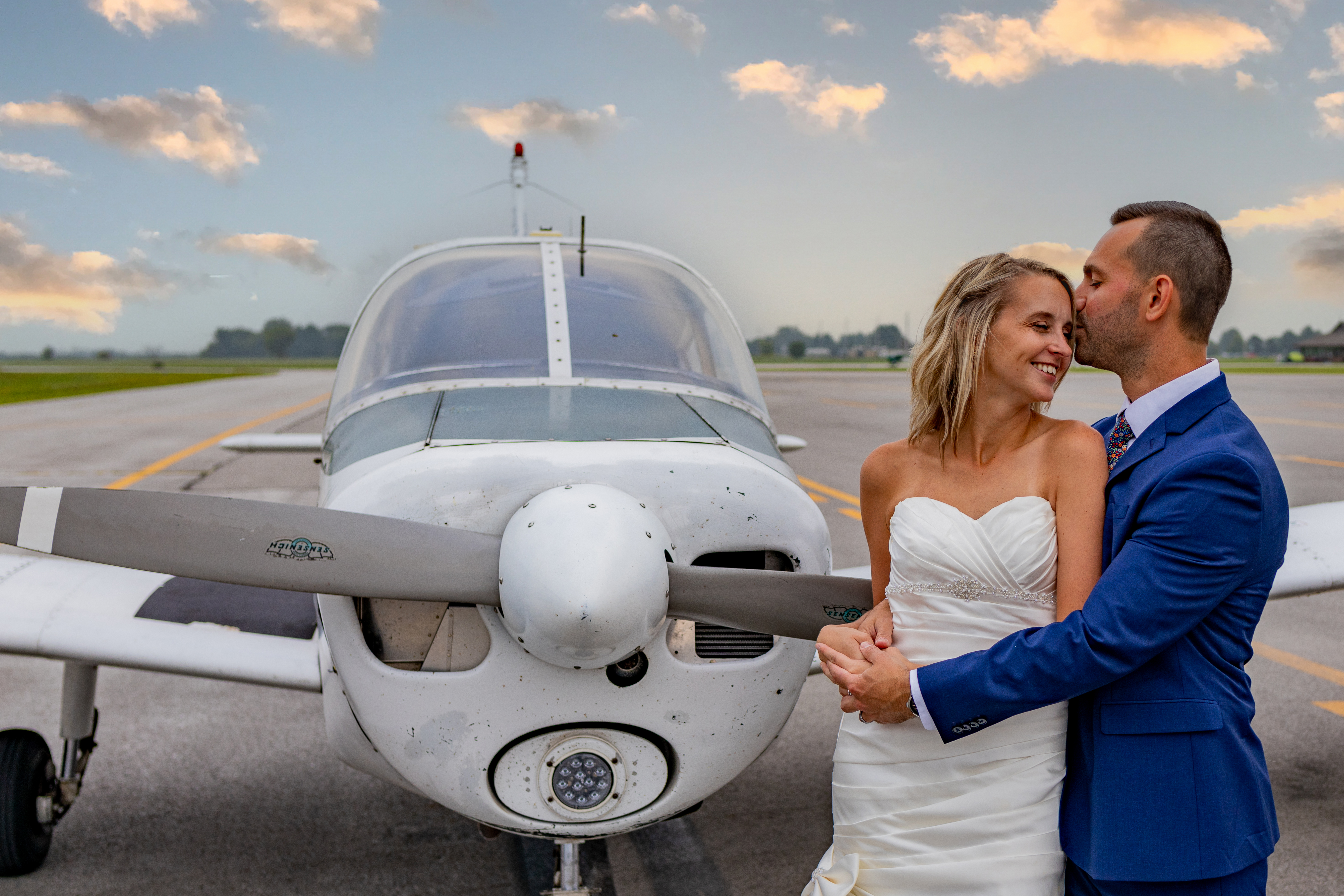 groom kissing bride on forehead in front of airplane